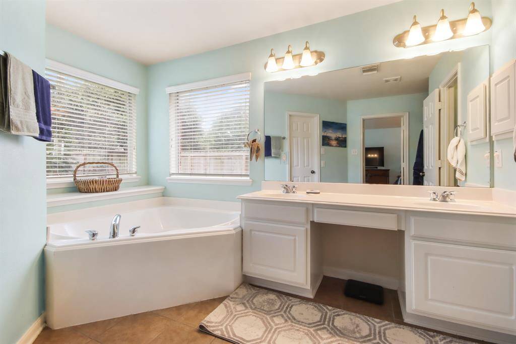 This bathroom features a spacious design with a large corner bathtub and a wide dual-sink vanity. The room is bright, thanks to ample natural light from two windows with blinds. The soft blue walls and modern lighting create a relaxing atmosphere, complemented by tile flooring and a convenient doorway leading to primary closet.