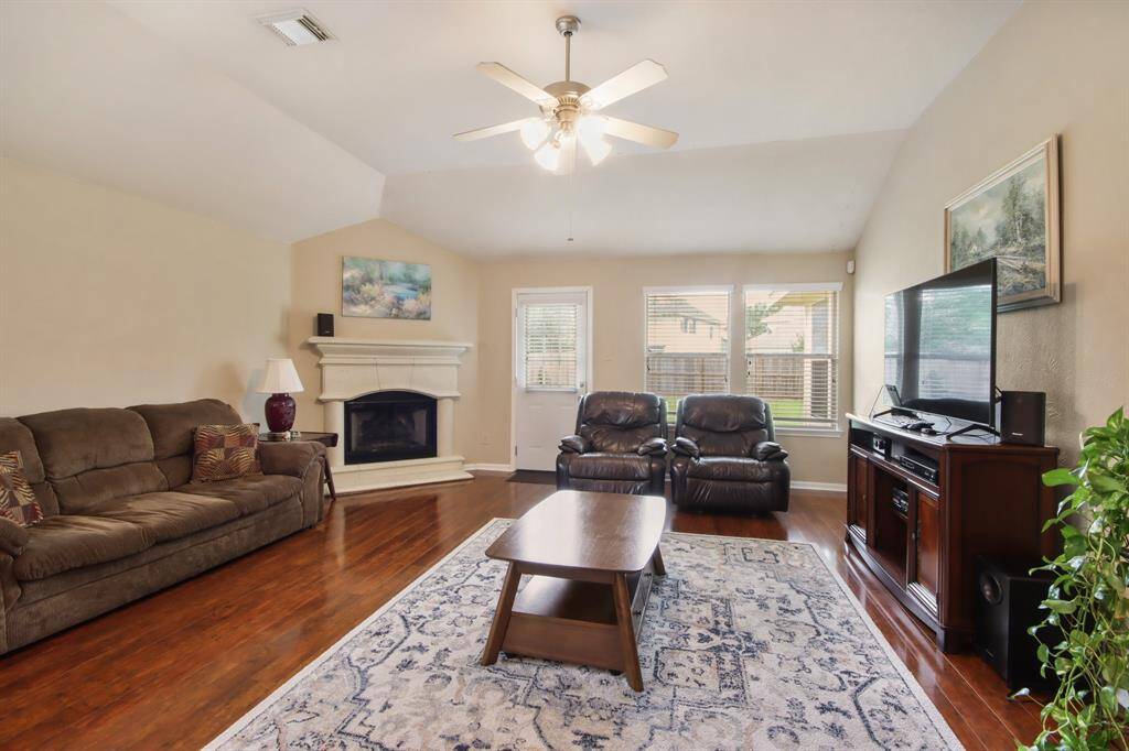 This spacious living room features hardwood floors, a cozy fireplace, and ample natural light from large windows. It includes accommodating square footage for comfortable seating and a ceiling fan, to create a welcoming space for relaxation and entertainment.