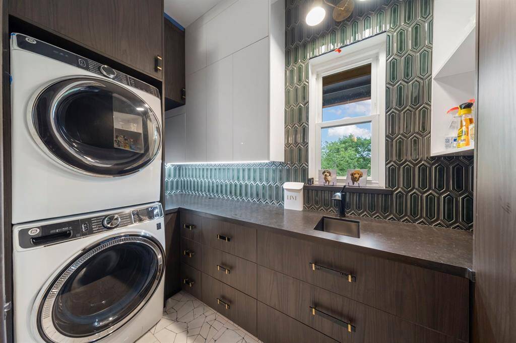 This modern laundry room features stacked appliances, sleek dark cabinetry with gold accents, bold geometric tile backsplashes, and a functional sink beneath a window for natural light.