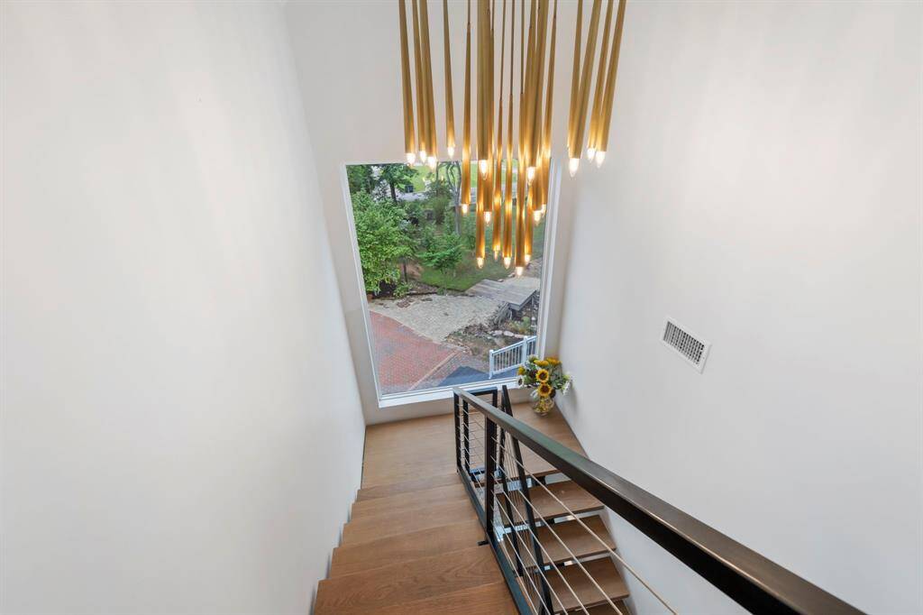 This staircase features clean lines, warm wood steps, a modern metal railing, and a dramatic cluster of gold pendant lights, all framed by a large picture window that floods the space with natural light.