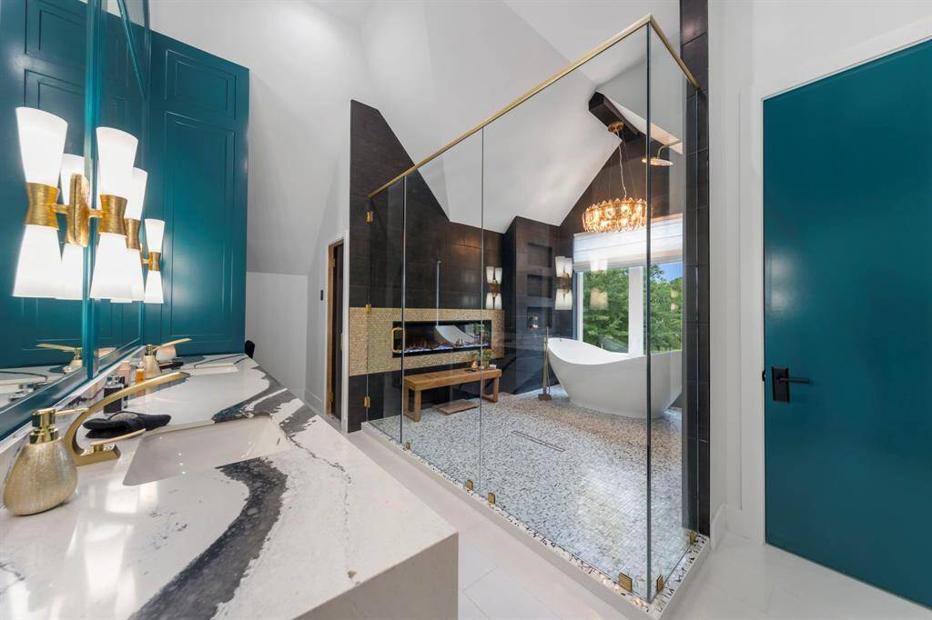 This luxurious bathroom features a bold teal accent wall, a striking vanity, a freestanding tub, and a glass-enclosed shower with a fireplace and chandelier for a spa-like atmosphere.