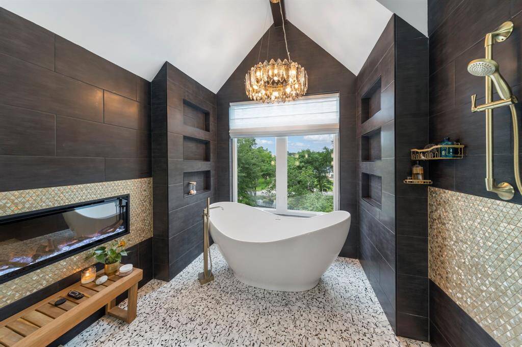 A sculptural soaking tub anchors this spa-like retreat, framed by dramatic dark tile, a modern fireplace, and a crystal chandelier beneath a vaulted ceiling with a view.