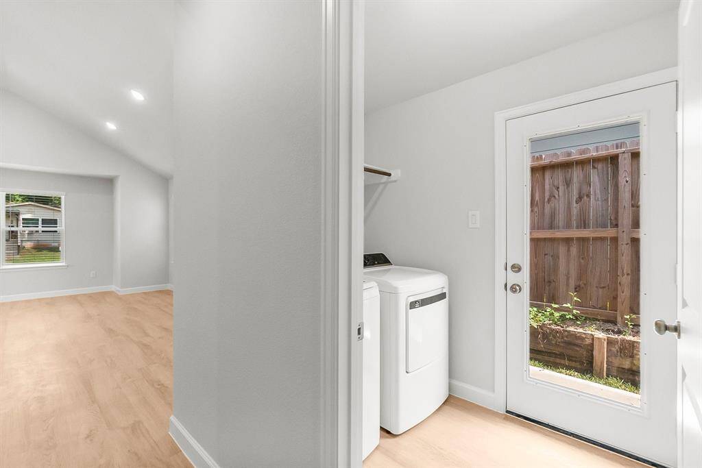 Bright, modern laundry area with washer and dryer, plus a door to the fenced backyard. Adjacent is a sunlit room featuring light wood flooring.