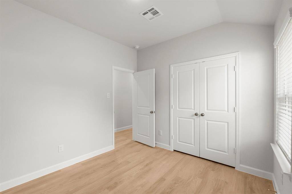 Bright and inviting bedroom featuring light wood flooring, crisp white walls, and a spacious double-door closet. Enjoy abundant natural light through the window with blinds.