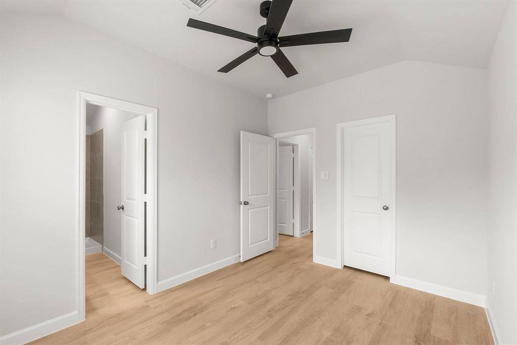 This room features light wood flooring, neutral walls, and a modern black ceiling fan. It includes two doors, one leading to a bathroom with tiled walls, and another to a hallway, offering a clean, contemporary feel.