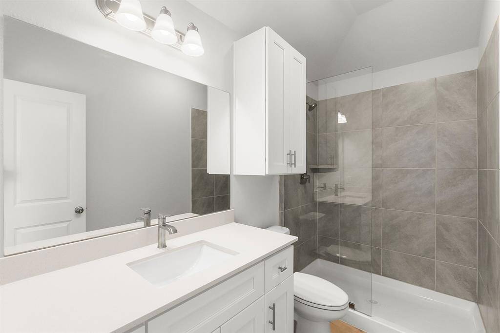 This modern bathroom features a sleek design with a large mirror, white cabinetry, and a glass-enclosed shower. The neutral gray tiles add a contemporary touch, complemented by ample lighting. Perfect for a stylish, functional space.