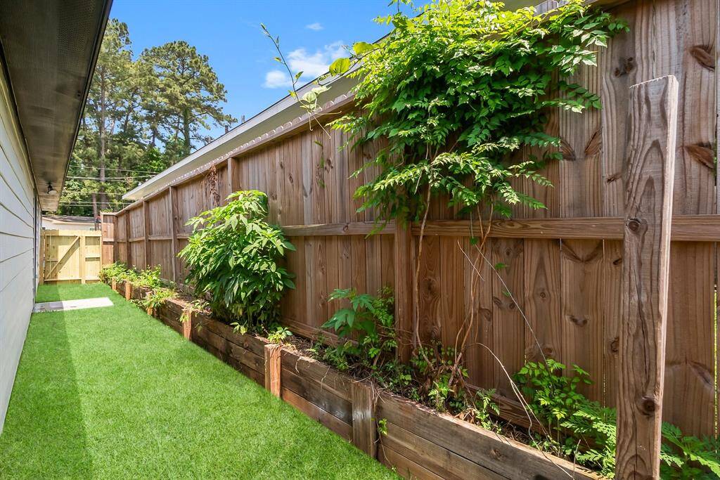 This photo shows a narrow side yard with a wooden fence and lush green grass. There are raised garden beds along the fence filled with various plants, offering a cozy and private outdoor space.