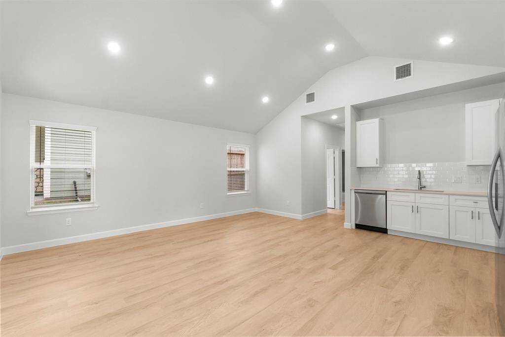 Bright and airy open-concept living area with high ceilings, recessed lighting, and light wood flooring. The modern kitchen boasts white cabinetry, stainless steel appliances, and a subway tile backsplash, enhanced by natural light from two windows.