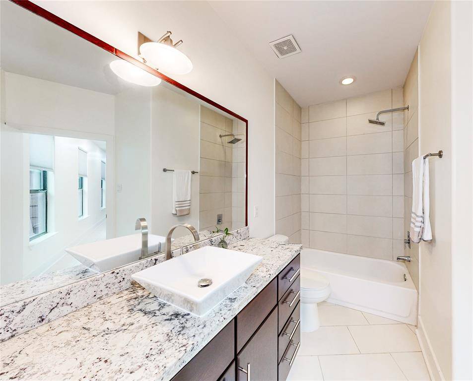 This bathroom features a modern design with a large granite countertop, vessel sink, and sleek fixtures. It includes a bathtub with a shower, tiled walls, and ample lighting, creating a bright and inviting space.