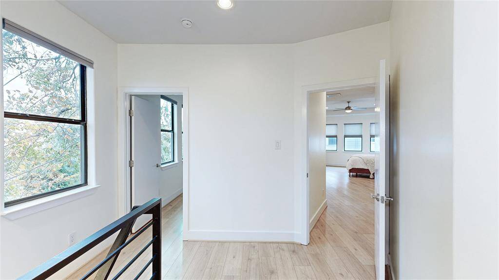 Bright hallway with light wood-look flooring leads to one of the secondary bedrooms and the Primary Bedroom. Features large windows, modern railing, and a neutral color palette.