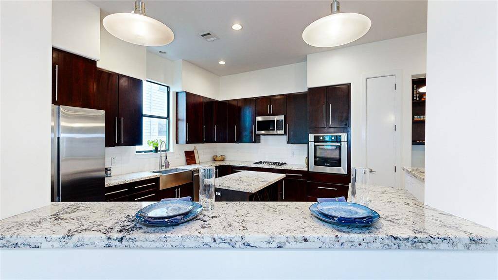 This modern kitchen features dark wood cabinets, stainless steel appliances, and a large central island. The space is brightened by pendant lighting and a window, offering a clean, contemporary look with ample counter space.