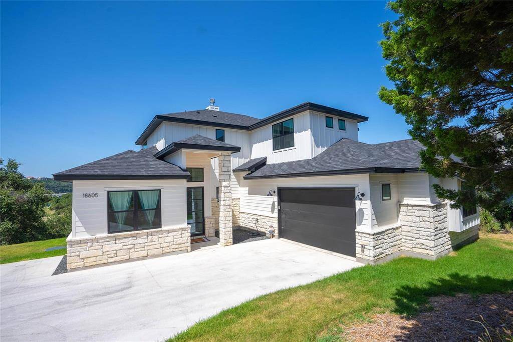 his modern two-story home features a sleek design with a combination of stone and siding exterior. It includes a spacious driveway, a two-car garage, and is surrounded by lush greenery.