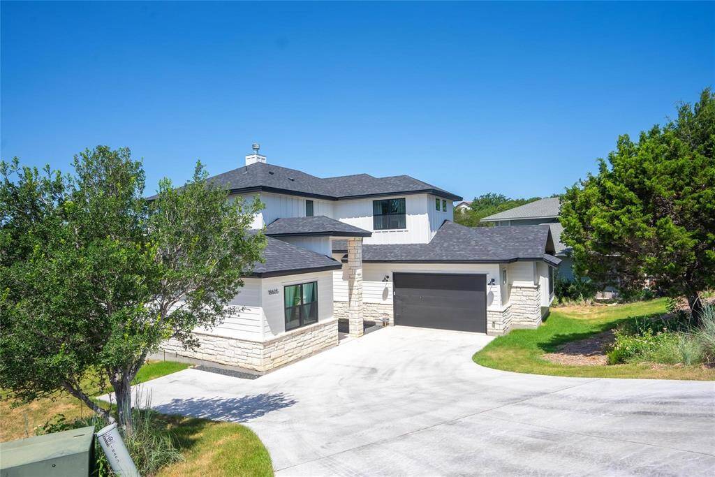 his modern two-story home features a sleek design with a combination of stone and siding exterior. It includes a spacious driveway, a two-car garage, and is surrounded by lush greenery.