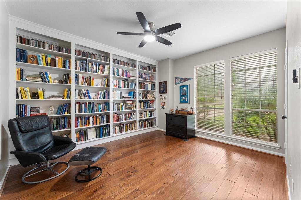 This bright and inviting den/office features a large built-in bookshelf filled with book, and two large windows with views of greenery. The space is enhanced by wood flooring and a ceiling fan, making it ideal for a home office or reading nook.