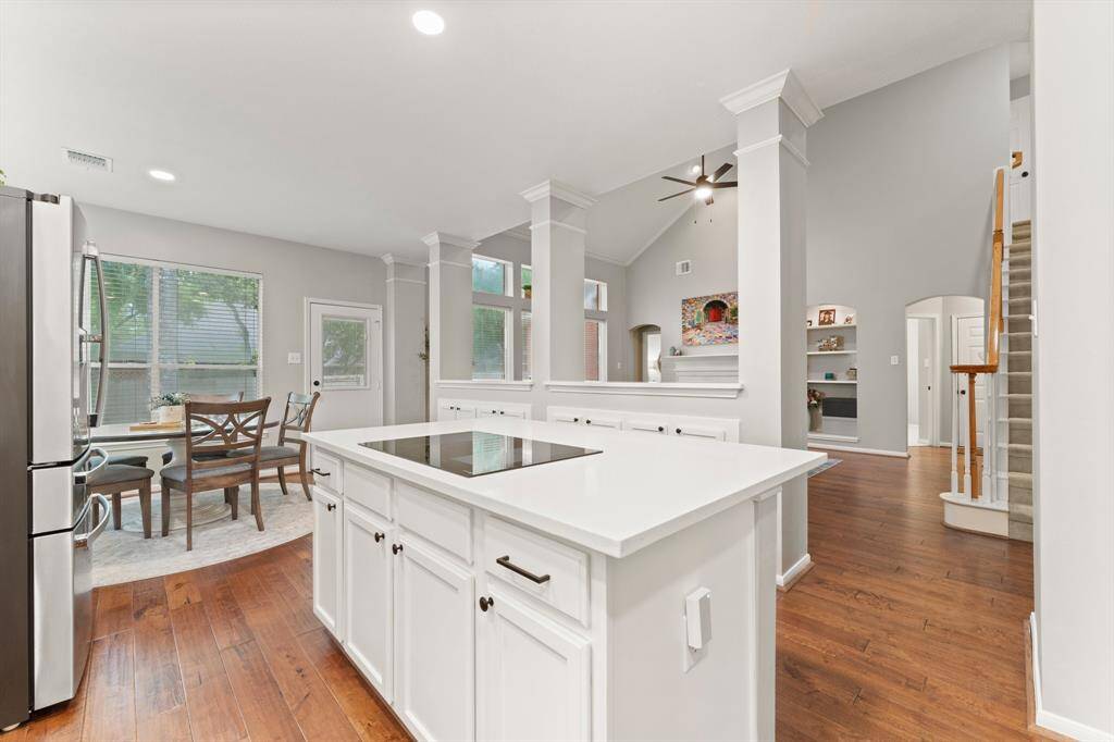 This open-concept kitchen and living area features a bright, airy design with high ceilings and neutral tones. It includes a modern kitchen island, stainless steel appliances, and hardwood floors, leading to a cozy dining nook and living space with built-in shelving and a fireplace.