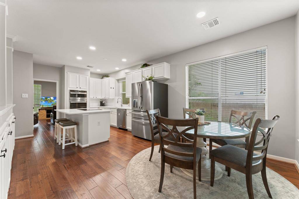 This photo showcases a modern kitchen with white cabinets, stainless steel appliances, and a central island with bar stools. The adjoining dining area features a round table with chairs, large windows, and hardwood flooring, creating a bright and open space.