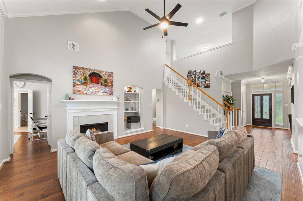 This inviting living room features high ceilings, a modern ceiling fan, and a cozy fireplace. The open layout includes a spacious sectional sofa and a prominent staircase leading to the upper floor. Natural light filters through large windows near the entrance, enhancing the warm wood flooring.