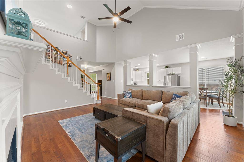 This photo showcases a spacious, open-concept living area with high ceilings and abundant natural light. The room features hardwood floors, a cozy sectional sofa, and a modern ceiling fan. An elegant staircase leads to the upper level, and the space seamlessly connects to a bright dining area and a contemporary kitchen with stainless steel appliances.