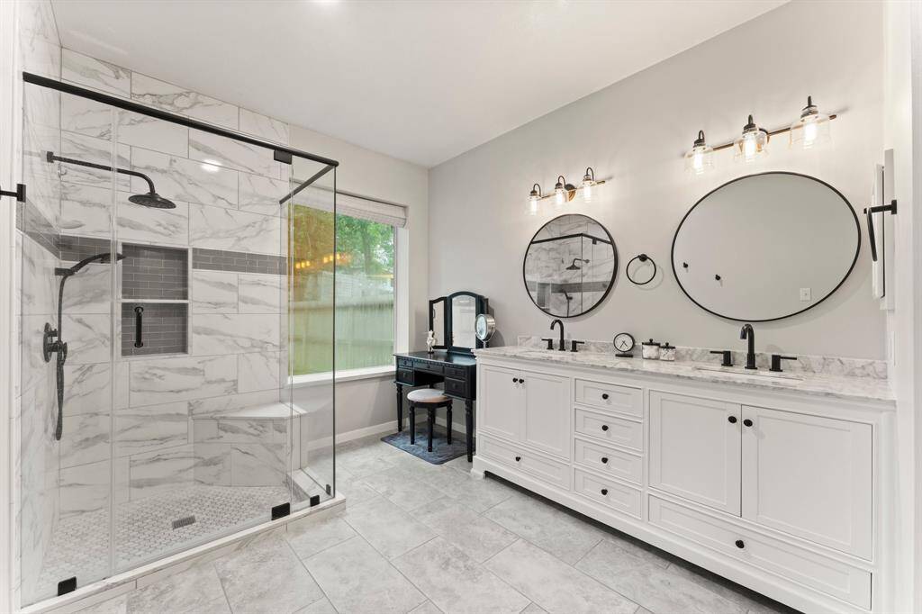 This modern bathroom features a spacious glass-enclosed shower with marble tile, a dual-sink vanity with marble countertops, and stylish black fixtures. It also includes a vanity table by a large window, providing ample natural light.