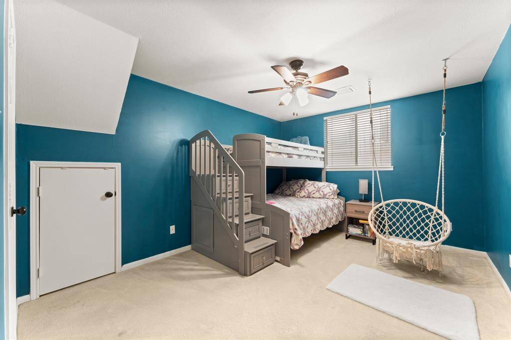 This cozy bedroom features vibrant blue walls, a ceiling fan, and a charming hanging chair. It has carpeted flooring, a window for natural light, and a small door for extra storage space, making it ideal for a child or guest room.