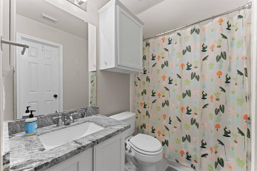 This is a small, modern bathroom featuring a granite countertop, white cabinetry, and a playful shower curtain with a botanical design. It includes a large mirror and neutral wall colors, providing a clean and bright appearance.