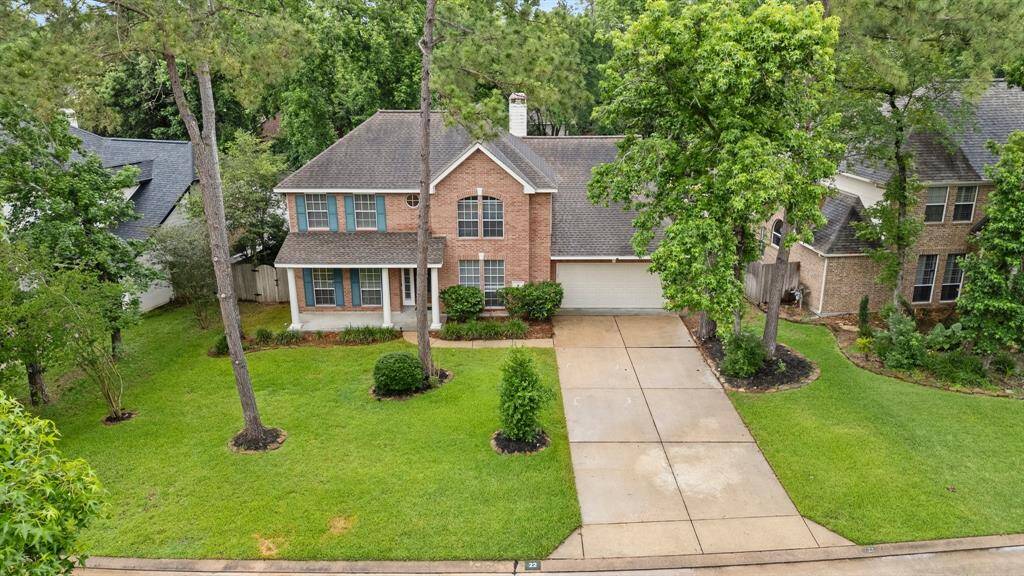 This charming two-story brick home features a classic facade with a covered front porch, surrounded by lush greenery and mature trees. It boasts a neatly manicured lawn and an attached three-car garage, offering a welcoming and serene atmosphere.