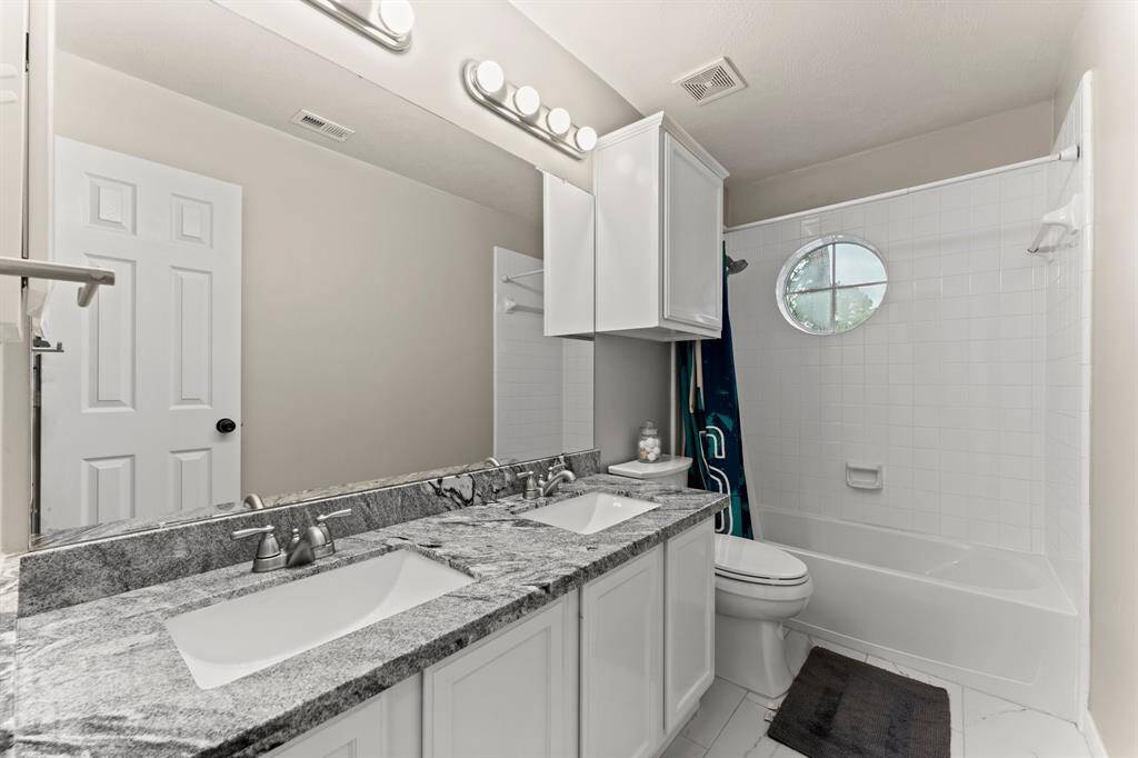 This bathroom features a dual-sink vanity with a granite countertop, a large mirror, and modern lighting. It includes a bathtub with a shower and a small circular window, creating a bright and functional space.