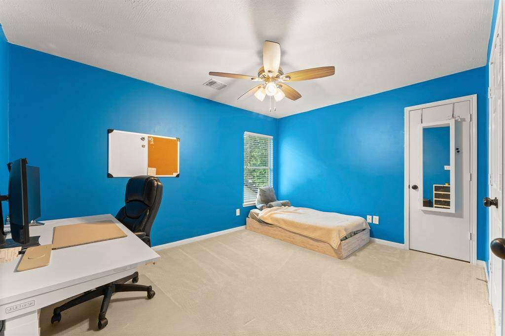 This bright room features vibrant blue walls, a ceiling fan, and neutral carpeting, ideal for a child's bedroom or home office. A window provides natural light, and a door has a full-length mirror.