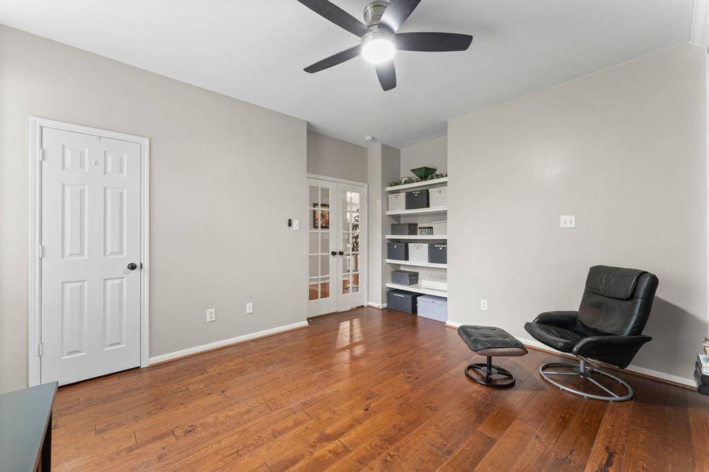 This den/office features hardwood floors, neutral walls, and a ceiling fan. It includes built-in shelving for storage, and a door with glass panels for natural light.