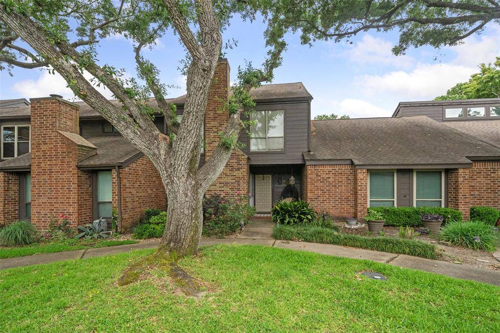 Charming two-story brick townhouse with a welcoming front entrance, surrounded by lush greenery and mature trees. The property features a well-maintained lawn and a spacious, inviting home.