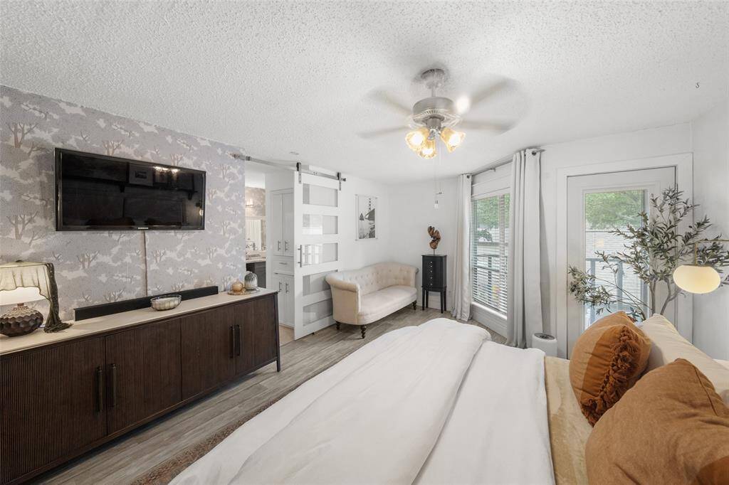 This primary bedroom features a cozy seating area, modern ceiling fan, and sliding barn door leading to an en-suite bathroom. Large windows and a glass door offer plenty of natural light .