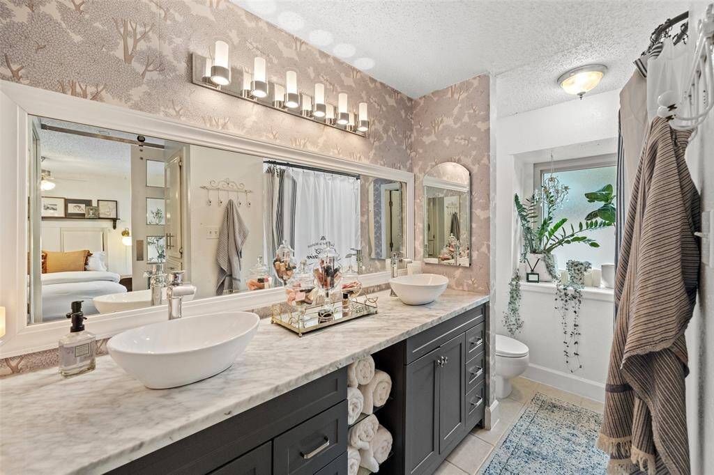 This elegant bathroom features a double vanity with vessel sinks and a marble countertop. The stylish wallpaper and large mirror add a touch of sophistication, while the well-placed lighting creates a warm ambiance. A window provides natural light.