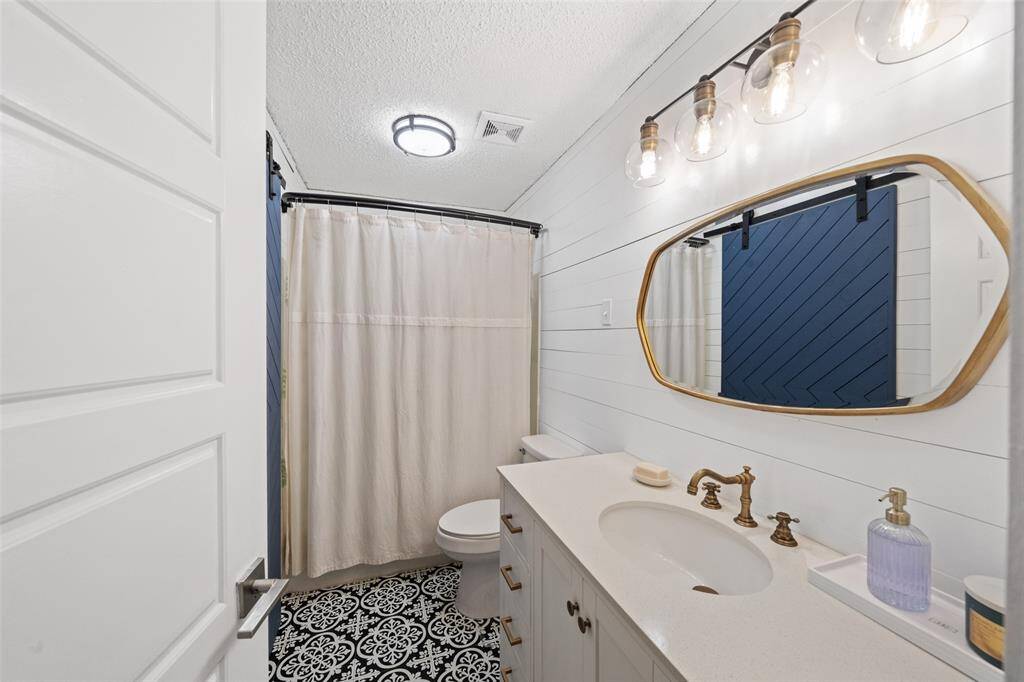 This bathroom features a modern design, a stylish vanity with brass fixtures, and patterned black-and-white floor tiles. The space is well-lit with a chic light fixture above a large mirror. completing the look.