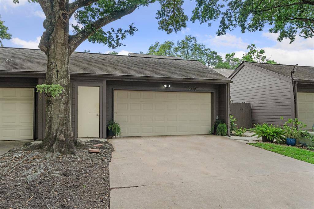 This photo shows a two-story townhouse with a clean, neutral exterior. It features a two-car garage and a driveway surrounded by mature trees and minimal landscaping .