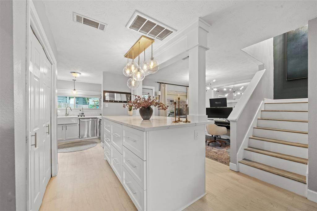 Bright and modern hallway with light wood floors, featuring a kitchen to the left with white cabinetry and stainless steel appliances. Elegant pendant lighting hangs above, and an open staircase leads to the upper level. A cozy office space is visible on the right.