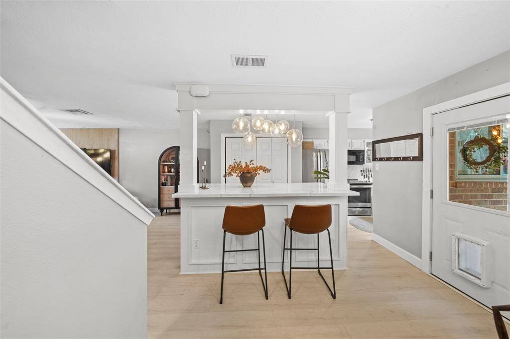 This photo showcases a modern kitchen with a breakfast bar. The open layout provides a view into the living area, emphasized by light wood flooring and contemporary lighting fixtures. A door with a pet flap adds convenience, and the space is bright and welcoming.