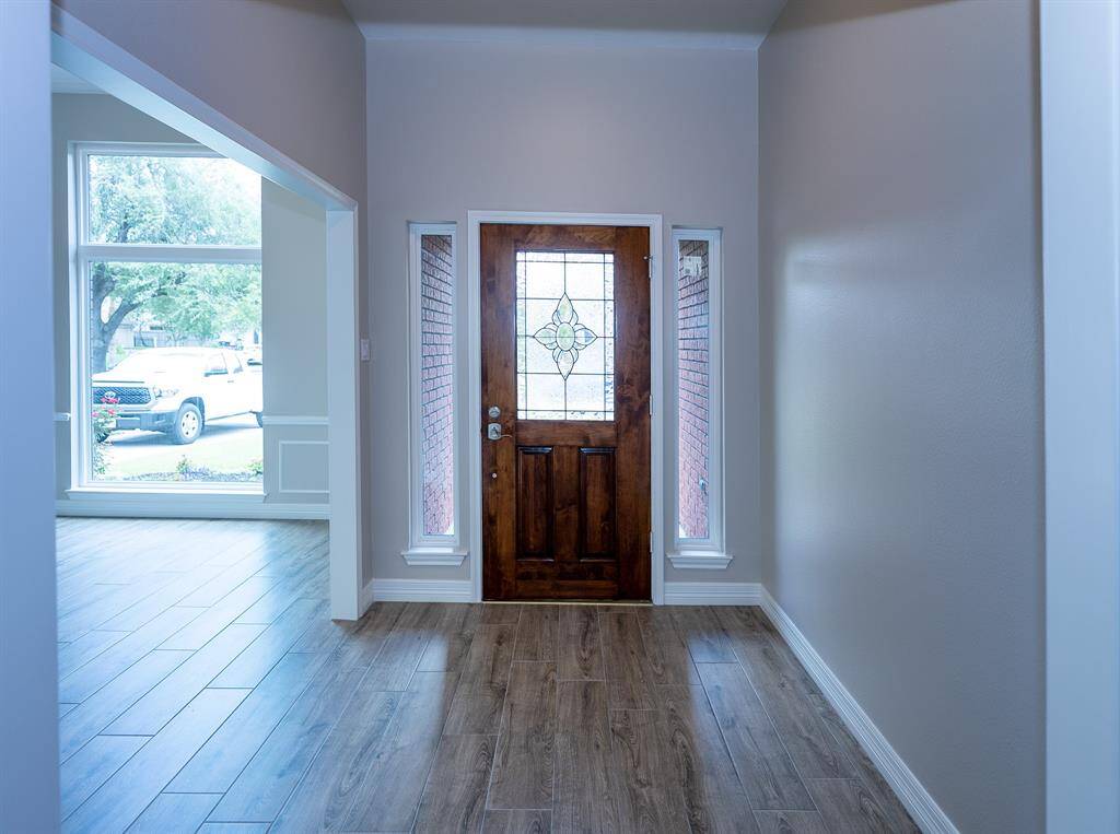 Inviting entryway with a wooden door featuring decorative glass panels. The space is bright with natural light from side windows, and has wood-look flooring leading into an open living area.