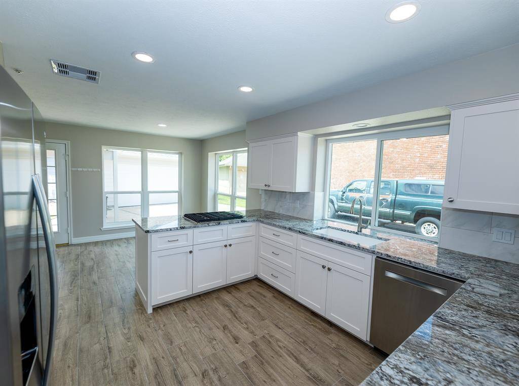 This kitchen features modern granite countertops, new white cabinetry, and stainless steel appliances. It has wood-style flooring and large windows for natural light, creating a bright and inviting space.