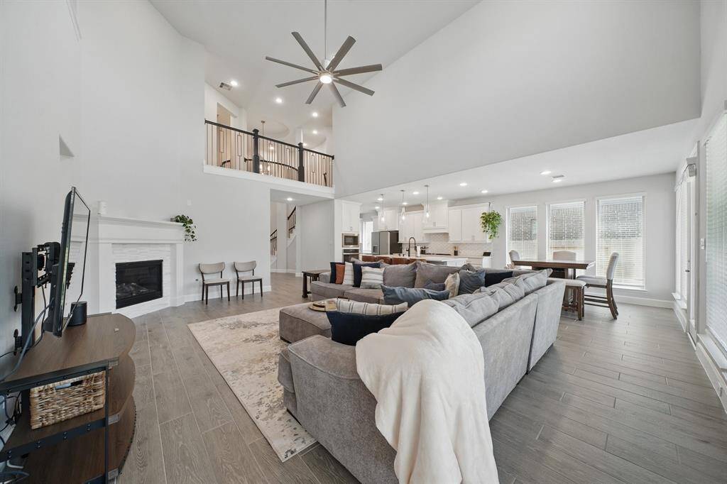 The living room showcases a two-story ceiling with balcony overlook, modern fan, and views into the open kitchen and breakfast area for effortless flow.