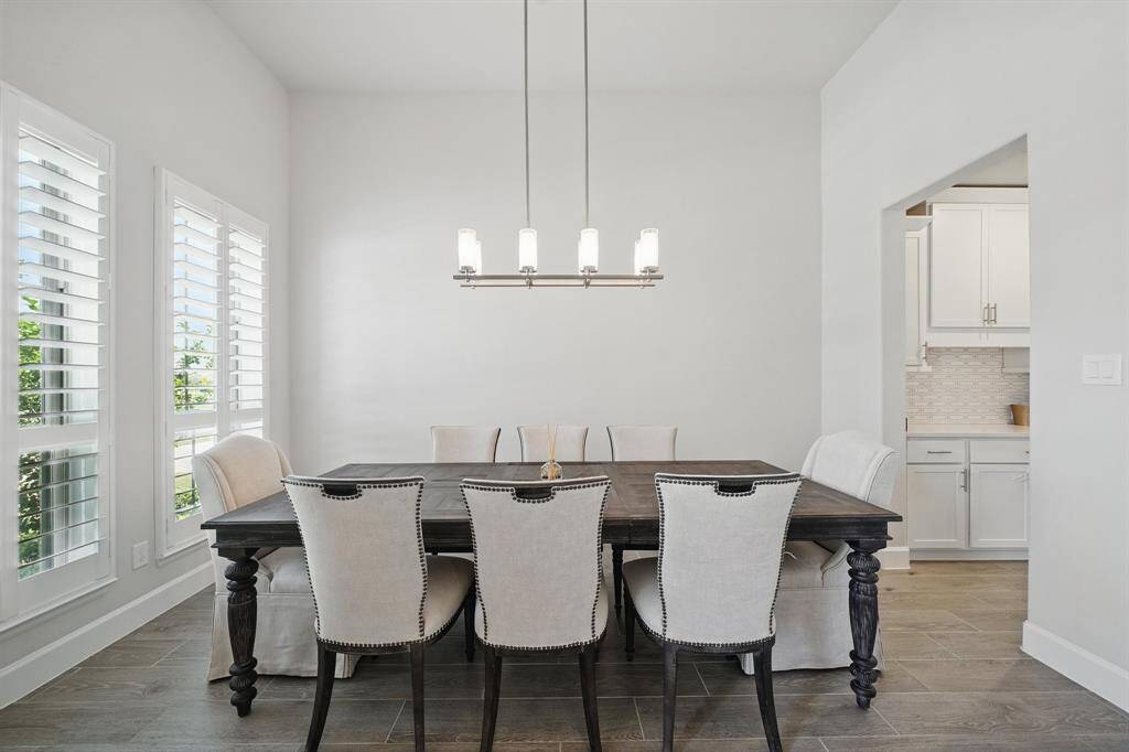 The formal dining room features tall ceilings, a sleek modern light fixture, and plantation shutters along three windows that bring in ample natural light.