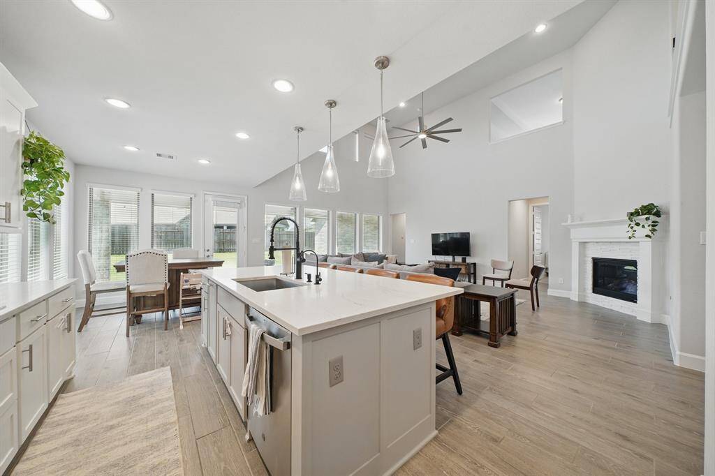 The open-concept layout connects the kitchen to the spacious living area, featuring a soaring ceiling, fireplace, and abundant natural light from walls of windows.
