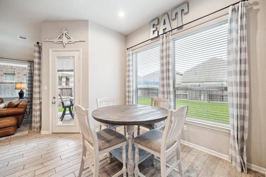 Bright breakfast nook with large windows, checkered curtains, and a rustic round table. The space opens to a cozy living area with a view of a well-kept backyard. Ideal for casual dining and entertaining.