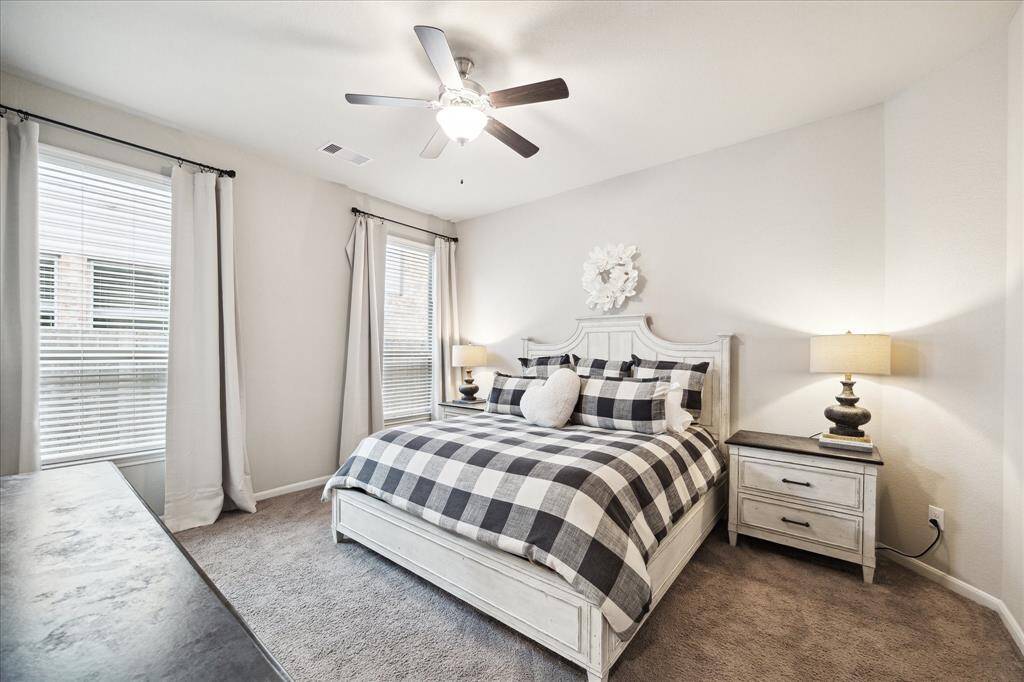 This inviting bedroom features a cozy, neutral color palette with ample natural light through two large windows, complemented by elegant drapes. The room includes a ceiling fan and plush carpeting creating a warm and relaxing atmosphere.