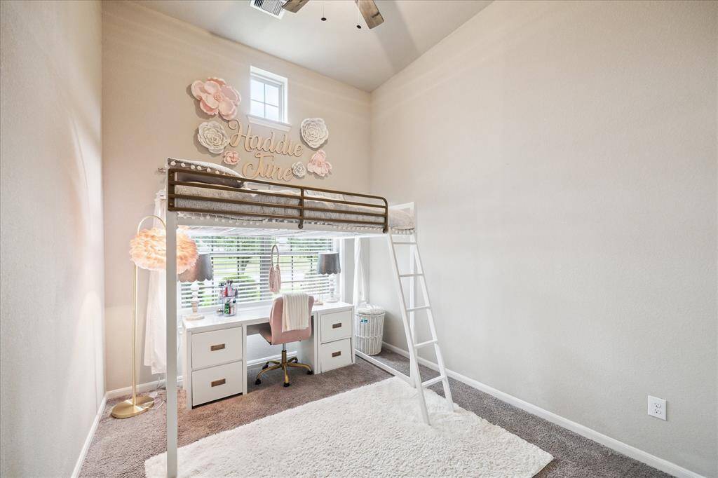 This is another bright bedroom with soft carpet flooring. The room has high ceilings and large window allowing natural light to filter in, creating a cheerful atmosphere.