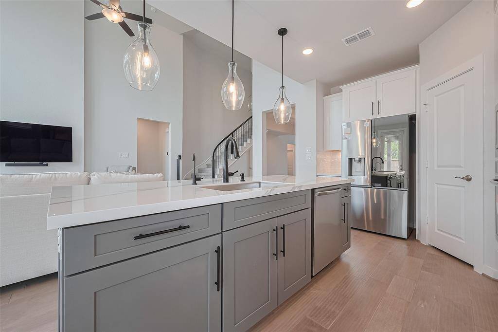 Modern kitchen with an open layout features sleek gray cabinetry, pendant lighting, and stainless steel appliances. The space flows into a living area with a wall-mounted TV and a view of the staircase, offering a bright and contemporary ambiance.