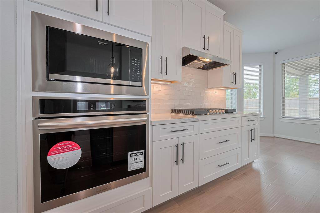This modern kitchen features sleek stainless steel appliances, including a built-in microwave and oven. It has elegant white cabinetry with black handles, a gas stovetop, and a stylish white tile backsplash. Natural light streams in from the adjacent windows, creating a bright and inviting space.