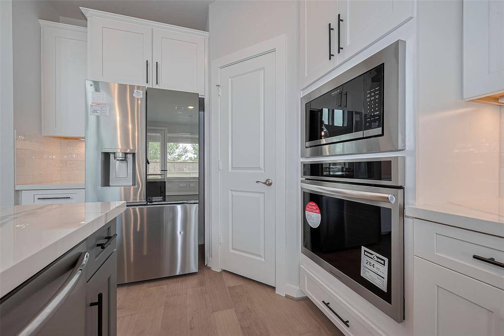 Features sleek stainless steel appliances, including a double wall oven and a large refrigerator. It has white cabinetry, a bright backsplash, and wood flooring for a clean, contemporary look.