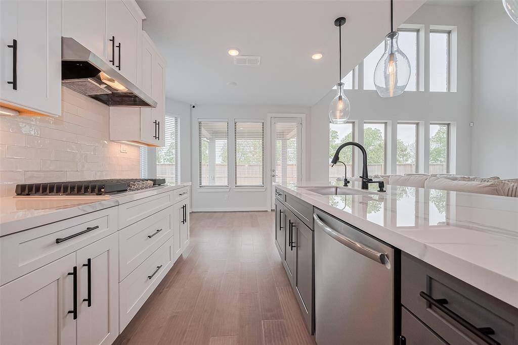 This modern kitchen features sleek white cabinetry, a spacious island with a sink, stainless steel appliances, and large windows that provide ample natural light. The open layout and high ceilings create an airy, inviting space.