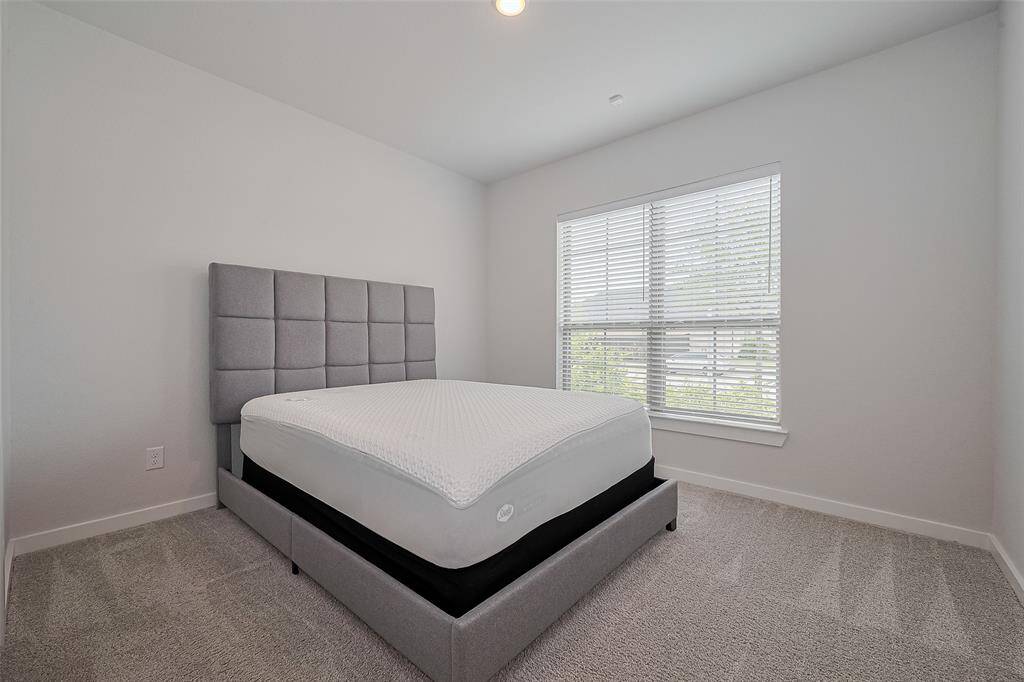 This bedroom features a modern design with a plush gray headboard, a comfortable bed, neutral walls, and carpeted flooring. A large window allows for ample natural light, creating a bright and inviting space.
