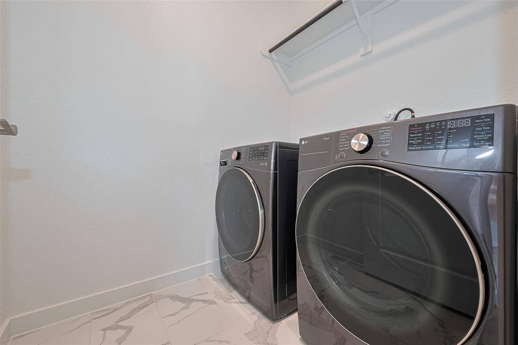 This photo shows a modern laundry room featuring a sleek washer and dryer set against a light-colored wall. The room has marble-patterned flooring and a white shelf for storage above the appliances.